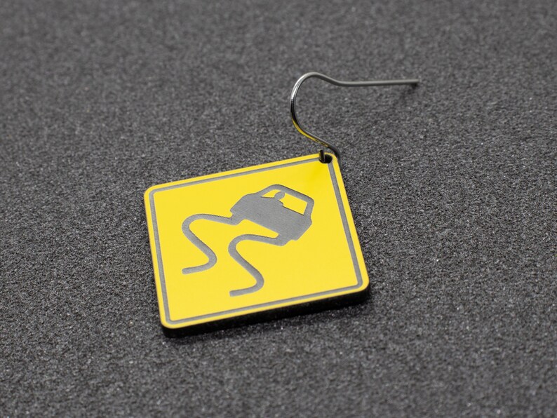 May include: A yellow and black car warning sign earring. The sign depicts a car skidding on a slippery road.