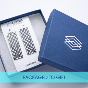 K&ouml;nnte beinhalten: Schwarze, lasergeschnittene Ohrringe in einer marineblauen Geschenkbox. Die Ohrringe haben ein geometrisches Gittermuster. Die Box hat ein wei&szlig;es Logo und den Schriftzug "PACKAGED TO GIFT".