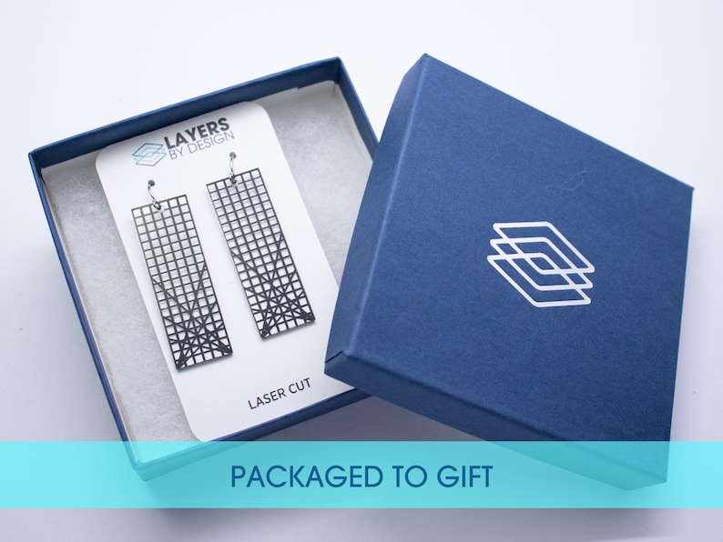K&ouml;nnte beinhalten: Schwarze, lasergeschnittene Ohrringe in einer marineblauen Geschenkbox. Die Ohrringe haben ein geometrisches Gittermuster. Die Box hat ein wei&szlig;es Logo und den Schriftzug "PACKAGED TO GIFT".