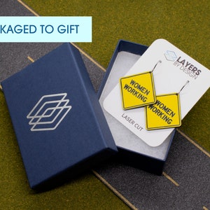 K&ouml;nnte beinhalten: Ein Paar gelbe und schwarze "Women Working"-Schildohrringe in einer blauen Geschenkbox. Die Ohrringe sind lasergeschnitten und werden mit einer wei&szlig;en Karte mit der Aufschrift "Layers by Design" geliefert.