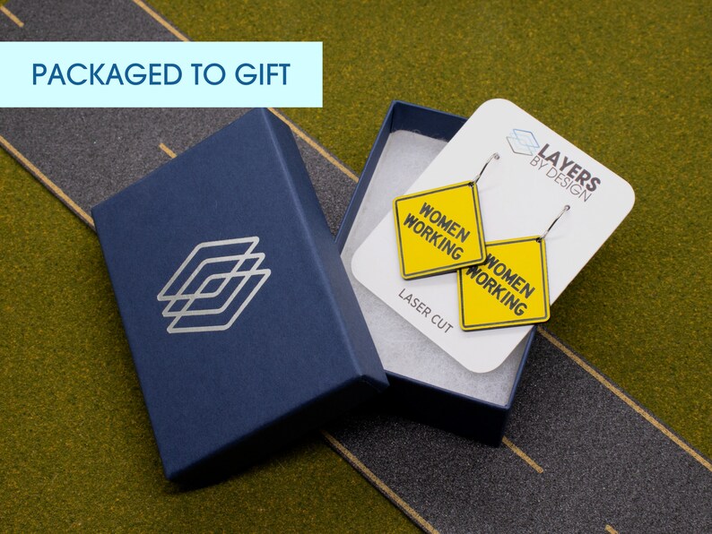 K&ouml;nnte beinhalten: Ein Paar gelbe und schwarze "Women Working"-Schildohrringe in einer blauen Geschenkbox. Die Ohrringe sind lasergeschnitten und werden mit einer wei&szlig;en Karte mit der Aufschrift "Layers by Design" geliefert.