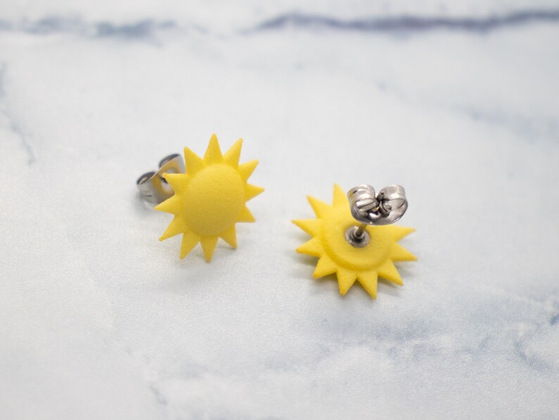 May include: A pair of yellow sun earrings with silver posts. The earrings are shaped like a sun with a starburst design.