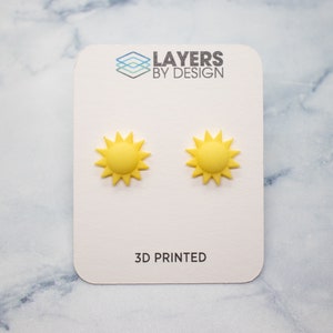 May include: A pair of yellow 3D printed sun earrings on a white card with the text "LAYERS BY DESIGN" and "3D PRINTED".