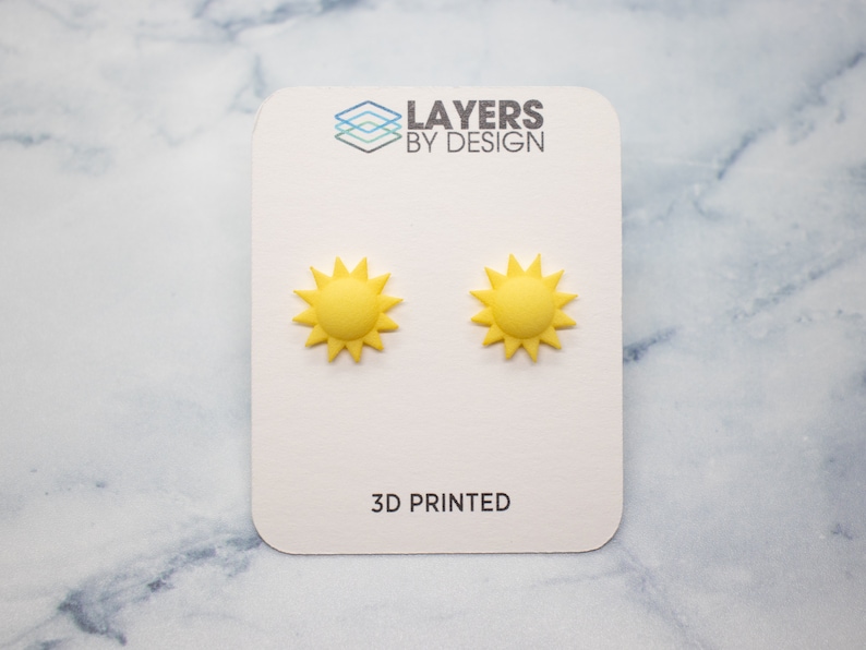 May include: A pair of yellow 3D printed sun earrings on a white card with the text "LAYERS BY DESIGN" and "3D PRINTED".