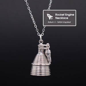 May include: A silver-colored rocket engine pendant necklace. The pendant is a detailed replica of a rocket engine, suspended from a delicate chain. The image includes a white rectangle with the text "Rocket Engine Necklace" and "Saturn V - NASA Inspired".