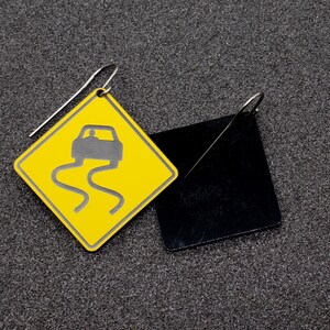 May include: A pair of earrings featuring a yellow diamond-shaped sign with a black car icon and a black square-shaped earring.