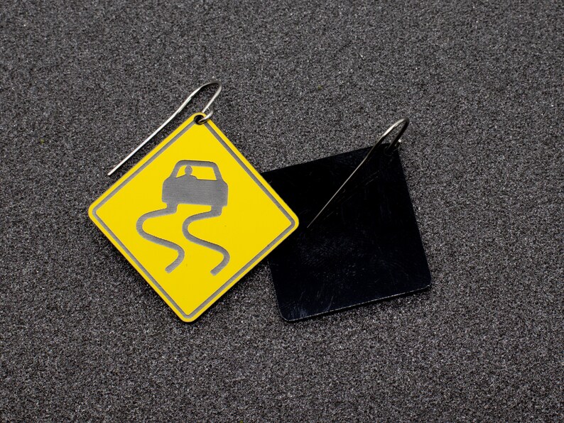 May include: A pair of earrings featuring a yellow diamond-shaped sign with a black car icon and a black square-shaped earring.