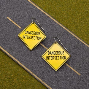 May include: A pair of yellow earrings shaped like a diamond road sign with the text "Dangerous Intersection". The earrings are on a gray surface that looks like a road with a yellow line down the middle.