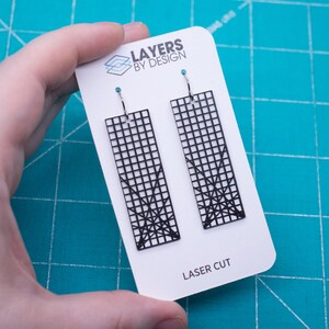 K&ouml;nnte beinhalten: Schwarze, lasergeschnittene Ohrringe mit einem geometrischen Gittermuster, pr&auml;sentiert auf einer wei&szlig;en Karte. Die Ohrringe haben eine rechteckige Form mit einem Gittermuster und einem dekorativen Design am unteren Rand. Auf der Karte steht "Layers by Design" und "Laser Cut".