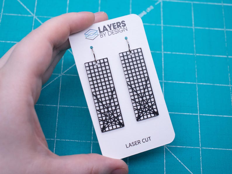 K&ouml;nnte beinhalten: Schwarze, lasergeschnittene Ohrringe mit einem geometrischen Gittermuster, pr&auml;sentiert auf einer wei&szlig;en Karte. Die Ohrringe haben eine rechteckige Form mit einem Gittermuster und einem dekorativen Design am unteren Rand. Auf der Karte steht "Layers by Design" und "Laser Cut".