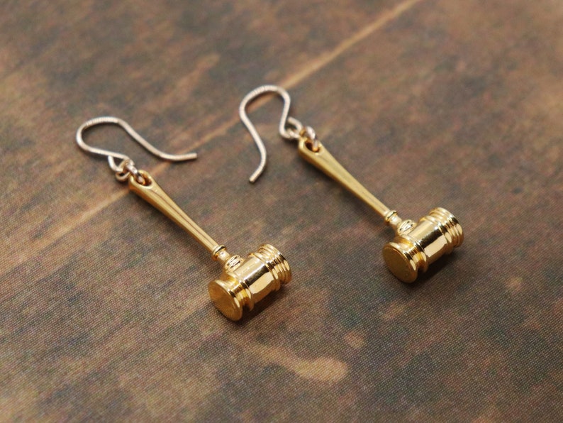 Puede incluir: Un par de pendientes de color dorado en forma de martillo. Los pendientes tienen un cierre de gancho simple.