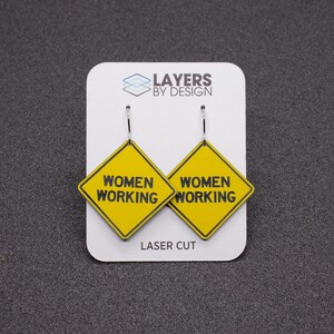 K&ouml;nnte beinhalten: Ein Paar gelbe 'Women Working'-Schild-Ohrringe mit schwarzem Text und silbernen Haken. Die Ohrringe werden auf einer wei&szlig;en Karte mit dem Text 'Layers by Design' und 'Laser Cut' pr&auml;sentiert.