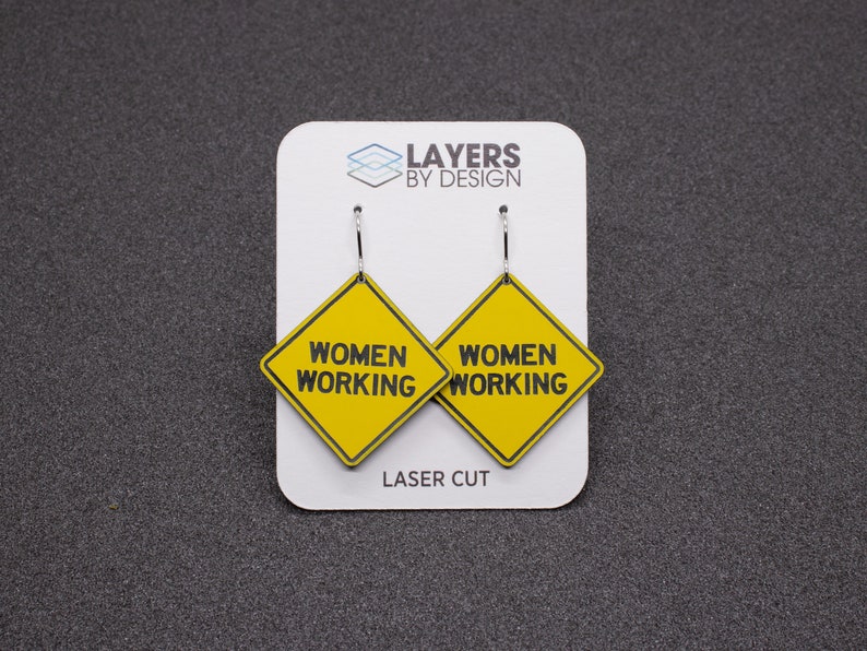 K&ouml;nnte beinhalten: Ein Paar gelbe 'Women Working'-Schild-Ohrringe mit schwarzem Text und silbernen Haken. Die Ohrringe werden auf einer wei&szlig;en Karte mit dem Text 'Layers by Design' und 'Laser Cut' pr&auml;sentiert.