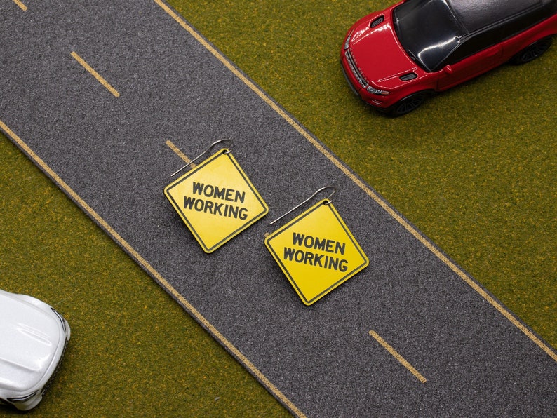 K&ouml;nnte beinhalten: Ein Paar gelbe und schwarze Ohrringe in Form von Verkehrsschildern mit dem Text "Women Working" darauf. Die Ohrringe befinden sich auf einer gr&uuml;nen Oberfl&auml;che mit einer grauen Stra&szlig;e im Hintergrund.