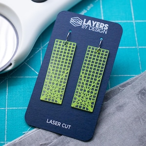 K&ouml;nnte beinhalten: Neongr&uuml;ne, lasergeschnittene Ohrringe mit geometrischem Gittermuster, pr&auml;sentiert auf einer marineblauen Karte. Die Karte tr&auml;gt den Text "Layers by Design" und "Laser Cut". Die Ohrringe sind rechteckig.
