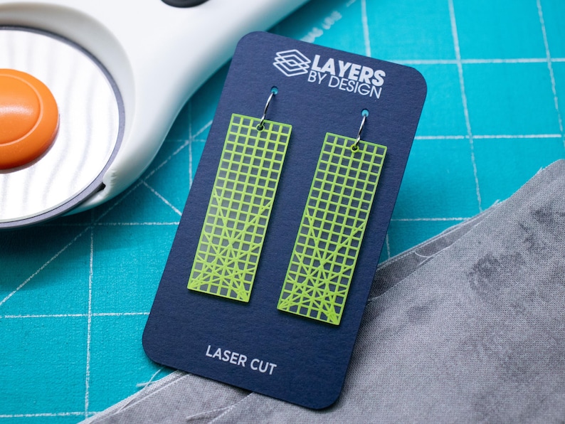 K&ouml;nnte beinhalten: Neongr&uuml;ne, lasergeschnittene Ohrringe mit geometrischem Gittermuster, pr&auml;sentiert auf einer marineblauen Karte. Die Karte tr&auml;gt den Text "Layers by Design" und "Laser Cut". Die Ohrringe sind rechteckig.
