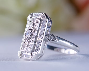 Puede incluir: Un anillo de plata con una piedra central rectangular rodeada de diamantes más pequeños. El anillo tiene un diseño delicado e intrincado.