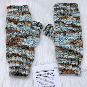 May include: A pair of knitted fingerless gloves in shades of blue, brown, and green, labelled "Autumns Embrace". The gloves are displayed on a white, fluffy surface. The label is attached to the gloves. The gloves are a cozy accessory.