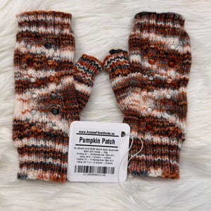 May include: A pair of fingerless gloves in a Pumpkin Patch colour scheme. The gloves are knitted with orange, brown, white, and grey yarn. The label reads "Pumpkin Patch" and includes knitting details.