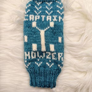 May include: Teal and white knitted dog sweater with the text "CAPTAIN HOWZER" and a tree design. The sweater features a ribbed cuff and hem, and a pattern of white chevrons.
