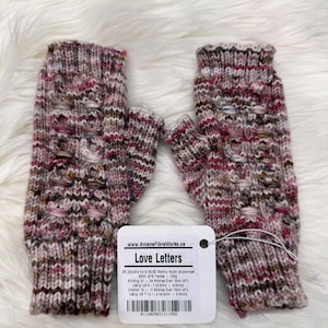 May include: Fingerless gloves in shades of pink, brown, and cream, with a textured knit design and small openings. The cuffs are ribbed. A tag attached to the gloves reads "Love Letters" and provides yarn details.