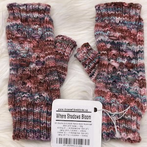 May include: A pair of hand-knitted fingerless gloves in pink, brown, and teal. The gloves have a textured pattern and are displayed on a fluffy white surface. A tag reads "Where Shadows Bloom".