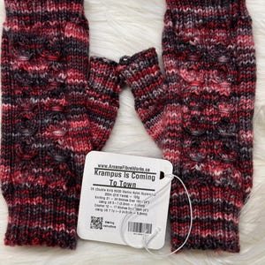 May include: Hand-knitted fingerless gloves in red, black, and grey. The gloves have a textured pattern and a ribbed cuff. A white tag with the text "Krampus Is Coming To Town" is attached. The gloves are made of wool.