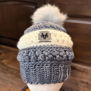 May include: A handmade, gray and cream-colored knit beanie with a faux fur pom-pom. The hat features a woven design and a small metal emblem with a logo. The beanie is displayed on a mannequin head.