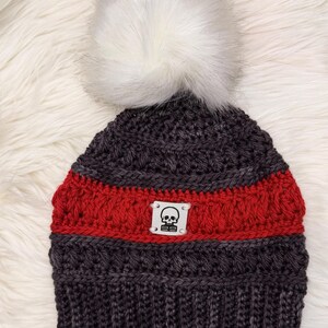 May include: A dark gray and red knit beanie with a white faux fur pom-pom. The hat features a red stripe and a small tag with a skull graphic. The beanie is displayed on a white, fluffy surface.