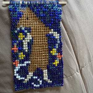 May include: A rectangular beaded wall hanging featuring a central gold-colored figure against a dark blue background. The design incorporates small white, yellow, and orange beads, suspended from a thin wooden dowel with a blue thread loop.