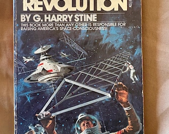 Può includere: Un libro tascabile intitolato The Third Industrial Revolution di G. Harry Stine. La copertina presenta un'illustrazione a tema spaziale con astronauti in tute arancioni e bianche che costruiscono una struttura metallica nello spazio.