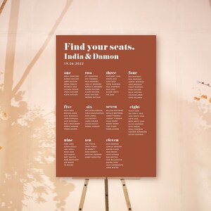 May include: A seating chart for a wedding reception with the names of guests listed under numbers one through eleven. The chart is printed on a terracotta-coloured card and is displayed on a wooden easel. The text at the top of the chart reads "Find your seats. India & Damon 19.06.2022".