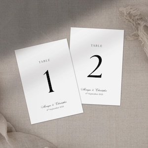 May include: Two white table number cards with black text. The cards read "TABLE" at the top, with the numbers "1" and "2" in a bold font. Below the numbers are names and a date. The cards are on a neutral-toned surface.