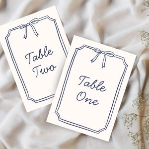 May include: Two ivory table cards with navy blue borders and bow accents. The cards read "Table Two" and "Table One" in elegant script. Suitable for weddings or events.