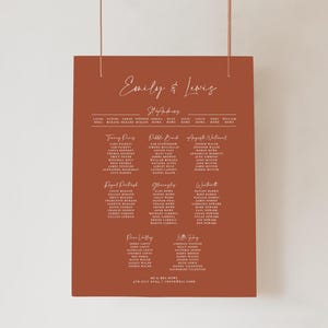 May include: A terracotta wedding seating plan with white calligraphy. The text reads "Emily & Lewis" at the top, followed by guest names organised by table. The date and venue are at the bottom. The chart is suspended by two thin strings.