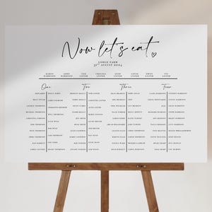 May include: A white wedding seating plan displayed on a wooden easel. The sign features the words "Now let's eat" in elegant calligraphy, with guest names listed by table. The date is 31st August 2024.