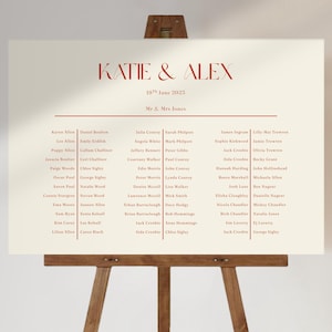 May include: A cream-coloured wedding seating plan displayed on a wooden easel. The chart features the names "KATIE & ALEX" in a red serif font, along with the date "10th June 2023". Guest names are listed below in two columns.