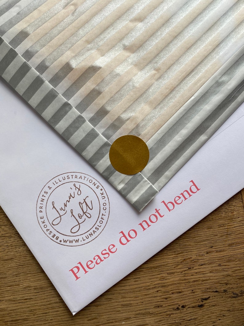 Puede incluir: Un sobre blanco con papel rayado plateado y blanco en su interior. El sobre tiene el texto "Please do not bend" impreso en rosa. Una pegatina circular dorada est&aacute; en el papel rayado.