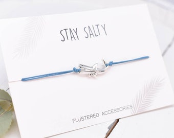 May include: A blue string bracelet with a silver shark charm. The bracelet has the text "STAY SALTY" and "FLUSTERED ACCESSORIES" printed on a white card.