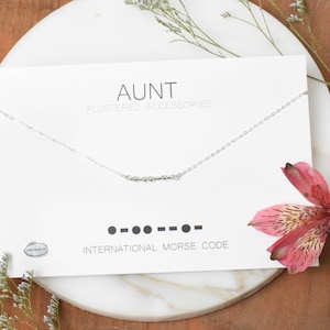 May include: A silver necklace with a small chain and a bar of beads spelling out "AUNT" in international Morse code. The necklace is displayed on a white card with the text "AUNT FLUSTERED ACCESSORIES" and "INTERNATIONAL MORSE CODE".