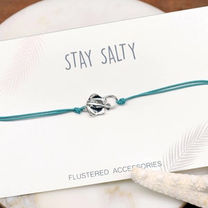 May include: A turquoise string bracelet with a silver charm that says "Stay Salty". The bracelet is on a white card with the words "Flustered Accessories" printed on it.