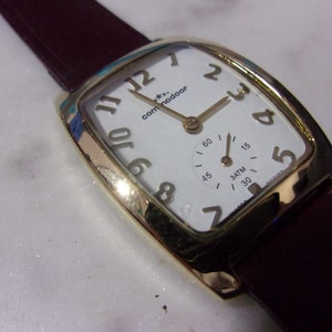 Peut inclure: Une montre-bracelet dorée avec un cadran blanc et un bracelet en cuir noir. Le cadran de la montre comporte de grands chiffres arabes noirs et un petit cadran auxiliaire à 3 heures. Le cadran de la montre porte également l'inscription "commodoor".
