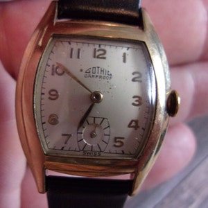 Peut inclure: Une montre-bracelet dorée avec un cadran blanc et un bracelet en cuir noir. Le cadran de la montre a des chiffres romains noirs pour les heures et un petit cadran des secondes. La montre est étiquetée "Gothil" et "Shockproof" sur le cadran.