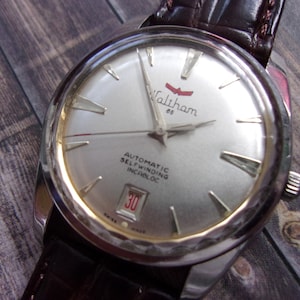 Può includere: Orologio da polso automatico Waltham 65 color argento con cinturino in pelle marrone. Il quadrante dell'orologio presenta lancette e indici argentati, un datario e il logo Waltham. L'orologio è in stile vintage.