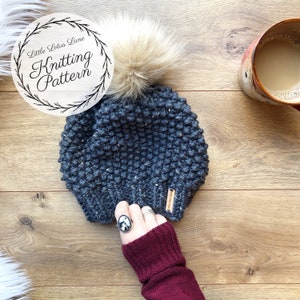 May include: A gray knitted beanie with a fluffy pom pom on top. The beanie has a brown leather tag with the text "Little Lotus Lane" on it. The beanie is on a wooden surface.