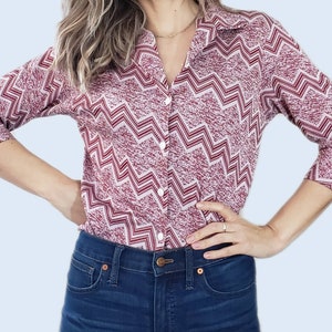 May include: A button-down shirt with a burgundy and white zigzag pattern. The shirt has a collar and long sleeves that end at the elbow. The shirt is paired with blue denim jeans.