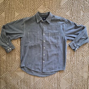 May include: A long-sleeve button-down shirt in a dark gray color with a textured fabric finish. The shirt features a classic collar, two chest pockets, and white buttons down the front placket and on the cuffs. A black GAP brand label is visible.