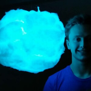 May include: A glowing, cloud-shaped light fixture emitting a bright blue hue. The fixture is suspended against a dark background. A person wearing a blue shirt is smiling in the right side of the image.