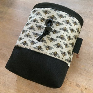 May include: A climbing chalk bag with a black base and top rim. The main body features a cream-colored fabric patterned with yellow and black bees. It has a black drawstring closure and a small black loop.