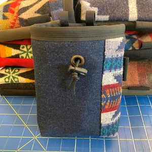 May include: A navy blue chalk bag with a patterned side panel featuring red, blue, and gray geometric designs. The bag has a drawstring closure and a black trim around the top. Several similar bags are stacked in the background.